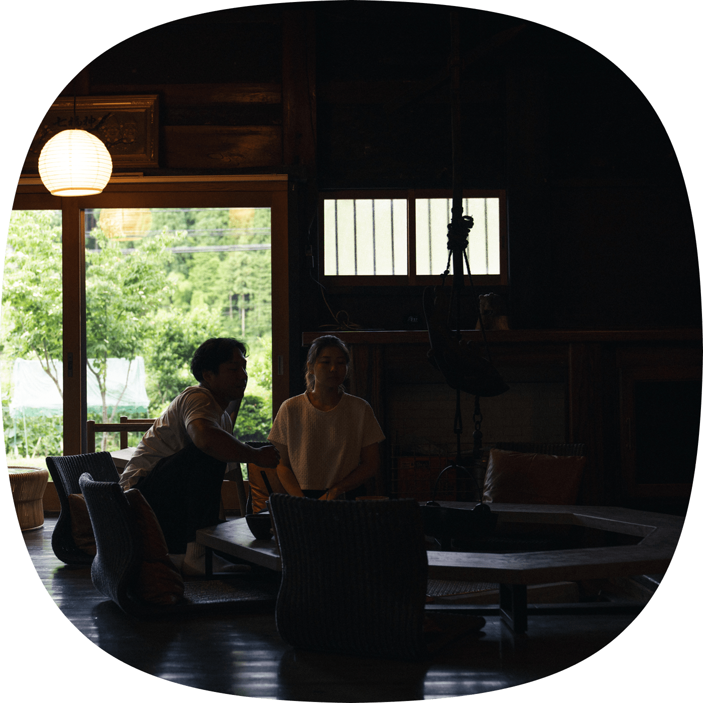 囲炉裏のある古民家で静かに語らう男女と、窓の外に広がる自然の風景