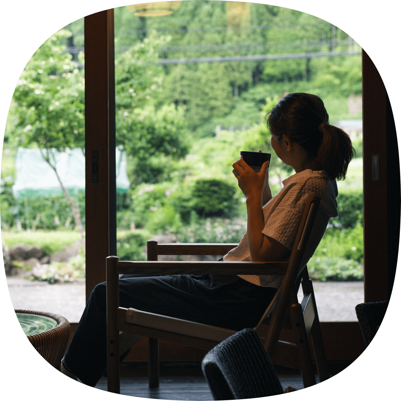 緑豊かな景色を眺めながら、椅子に座ってお茶を楽しむ女性の後ろ姿