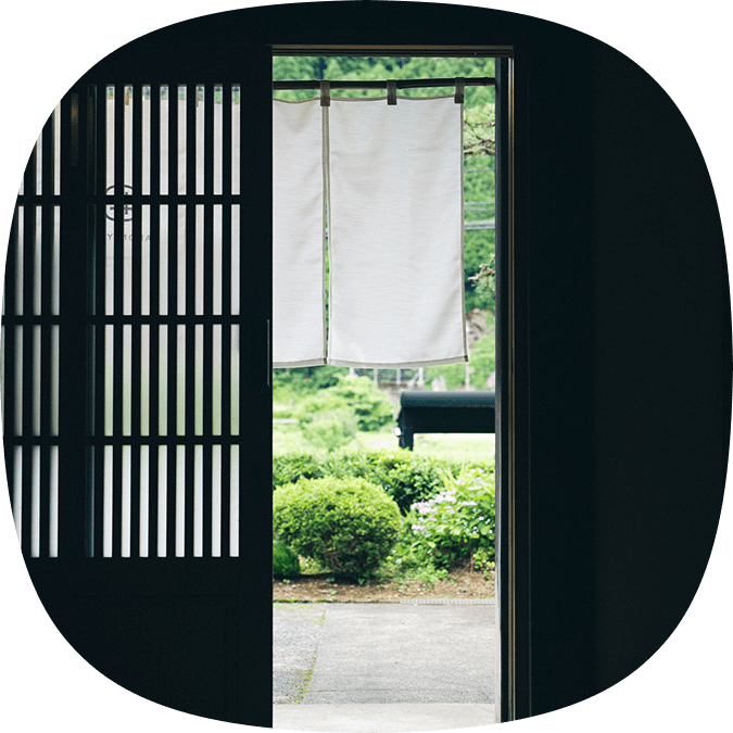 白い暖簾が揺れる古民家の玄関と、緑あふれる庭の景色