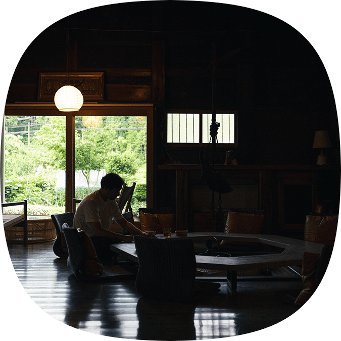 囲炉裏のそばでゆったりと過ごす男性と、窓の外に広がる緑の風景